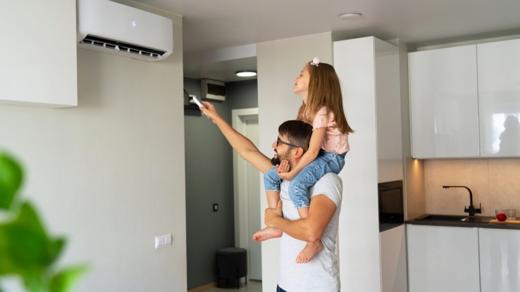 A man carries a young girl on his shoulders as she points a remote control at a wall-mounted air conditioner in a modern kitchen. ac repair beech mountain