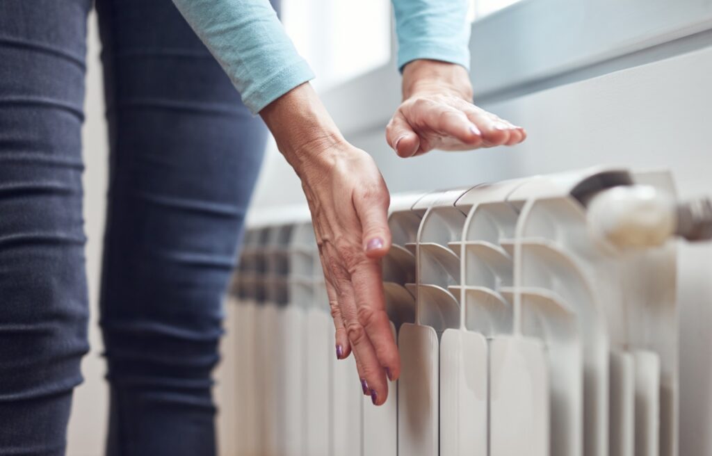 A person holds their hands over a white radiator, checking or feeling the heat, next to a window. Furnace Pilot Light