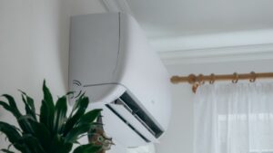 A wall-mounted air conditioner is installed near a window with sheer curtains; a green potted plant is visible in the foreground. why is my air conditioner so loud inside