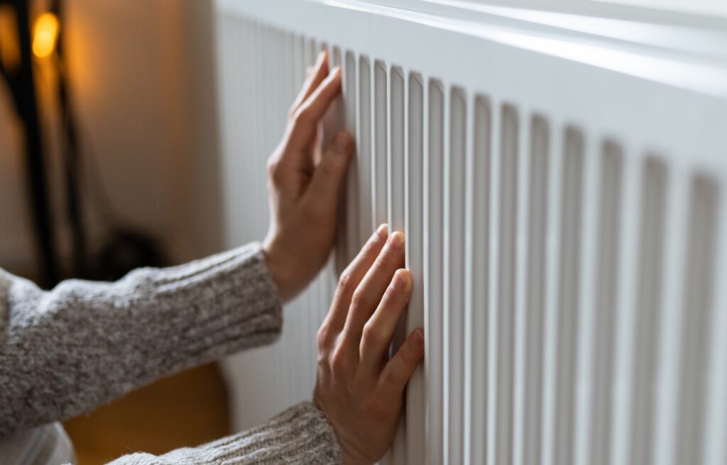 A person wearing a gray sweater places both hands on a white radiator, suggesting they are feeling its warmth.
