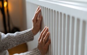 A person wearing a gray sweater places both hands on a white radiator, suggesting they are feeling its warmth. Heat Pump Not Heating