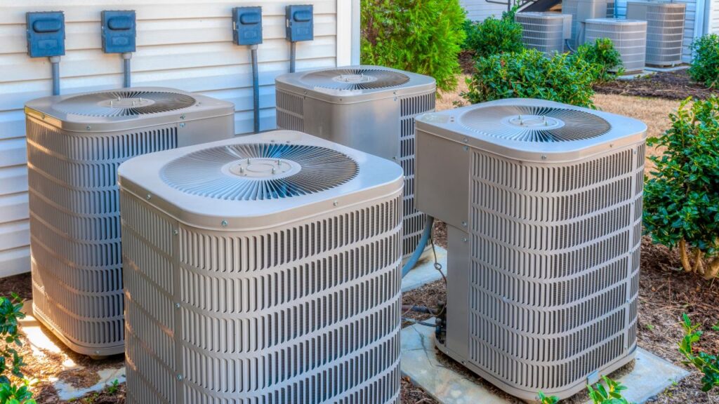 Four outdoor air conditioning units are installed side by side on concrete pads next to a building, surrounded by bushes and electrical panels.