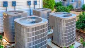 Four outdoor air conditioning units are installed side by side on concrete pads next to a building, surrounded by bushes and electrical panels. heating and ac repair near me Loud AC